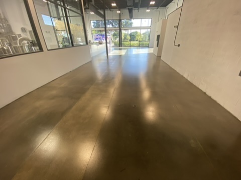 Glossy bronze-brown floor running the length of a narrow commercial space with glass partition walls on the left and a full-height storefront window at the far end