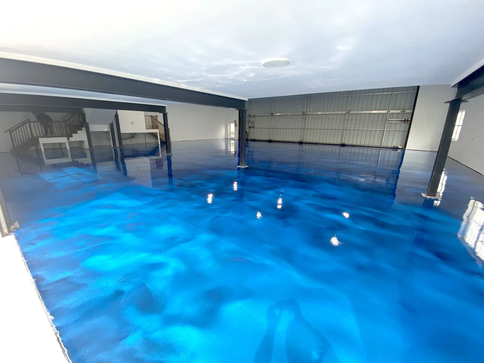 Wide open interior with a mirror-gloss electric blue metallic floor swirling under steel support posts, a white ceiling and a closed roll-up door in Estero, FL