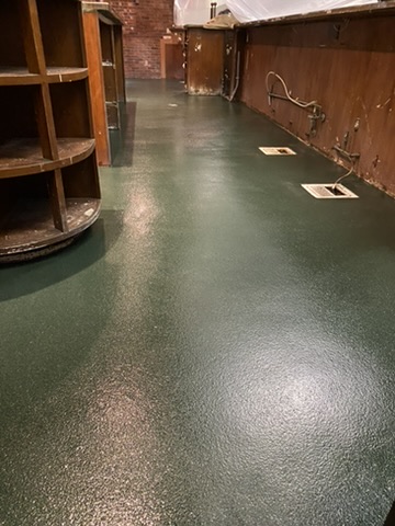 Dark green textured floor coating running the length of a narrow service corridor between a wooden shelving unit and a paneled wall in Estero, FL